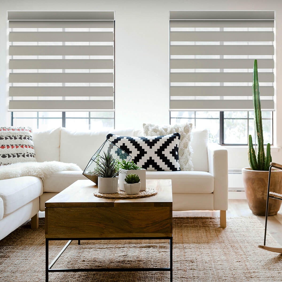 Beige zebra shades on two windows in a bright boho living room with a white sofa, patterned cushion and wooden coffee table