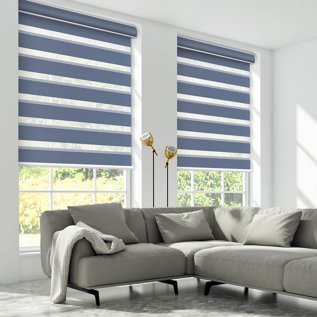 Blue zebra shades on two large windows above a grey sectional sofa with brass floor lamps in a bright living room