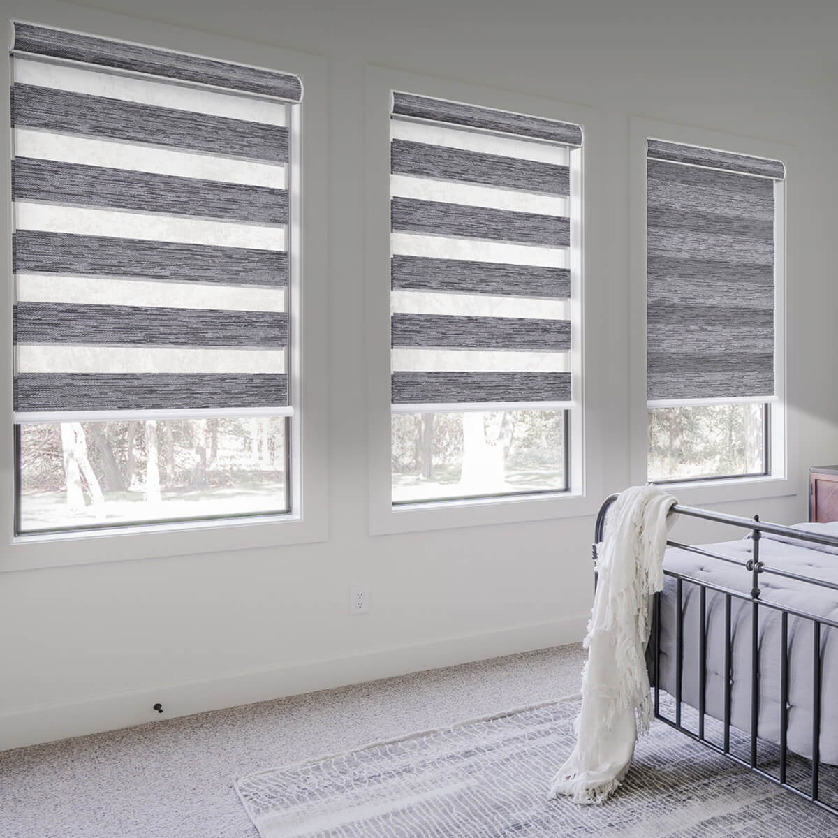 Light grey zebra shades on three bedroom windows beside a black metal bed with a white throw