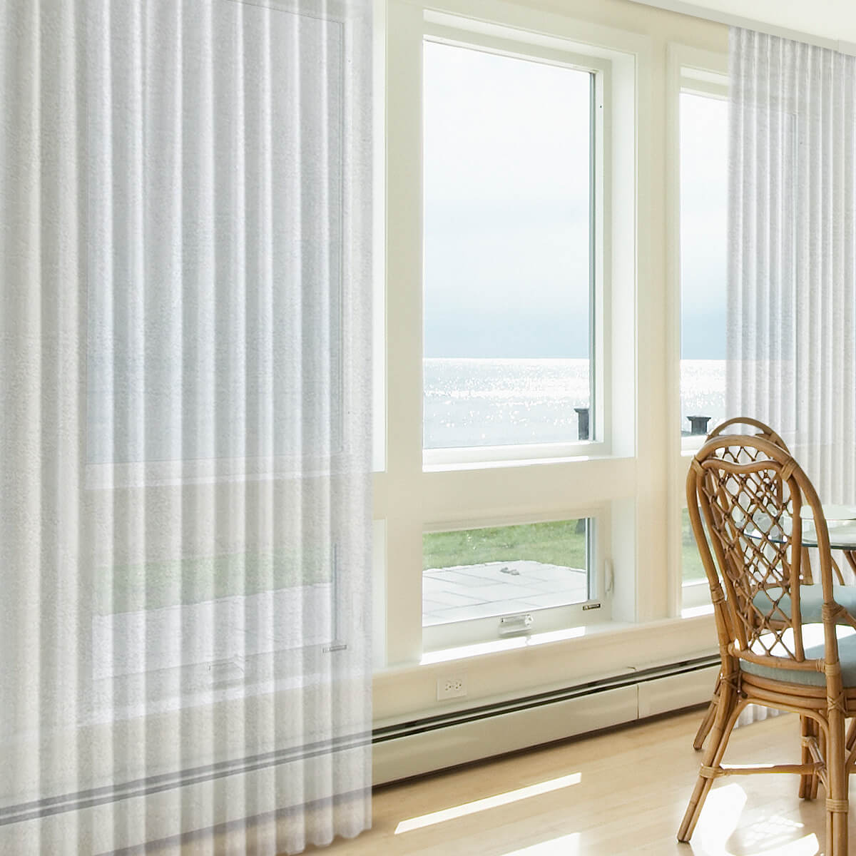 Sheer white motorized drapes drawn partly across a large glass opening with a sea view, beside a rattan chair