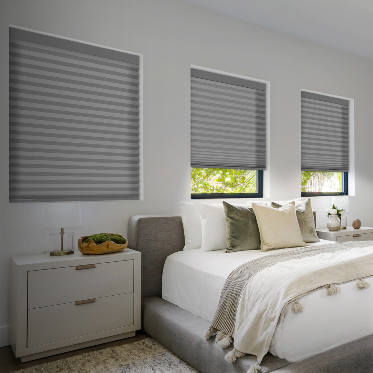 Ash grey light filtering cellular shades on three bedroom windows of different sizes above a bed with white linens